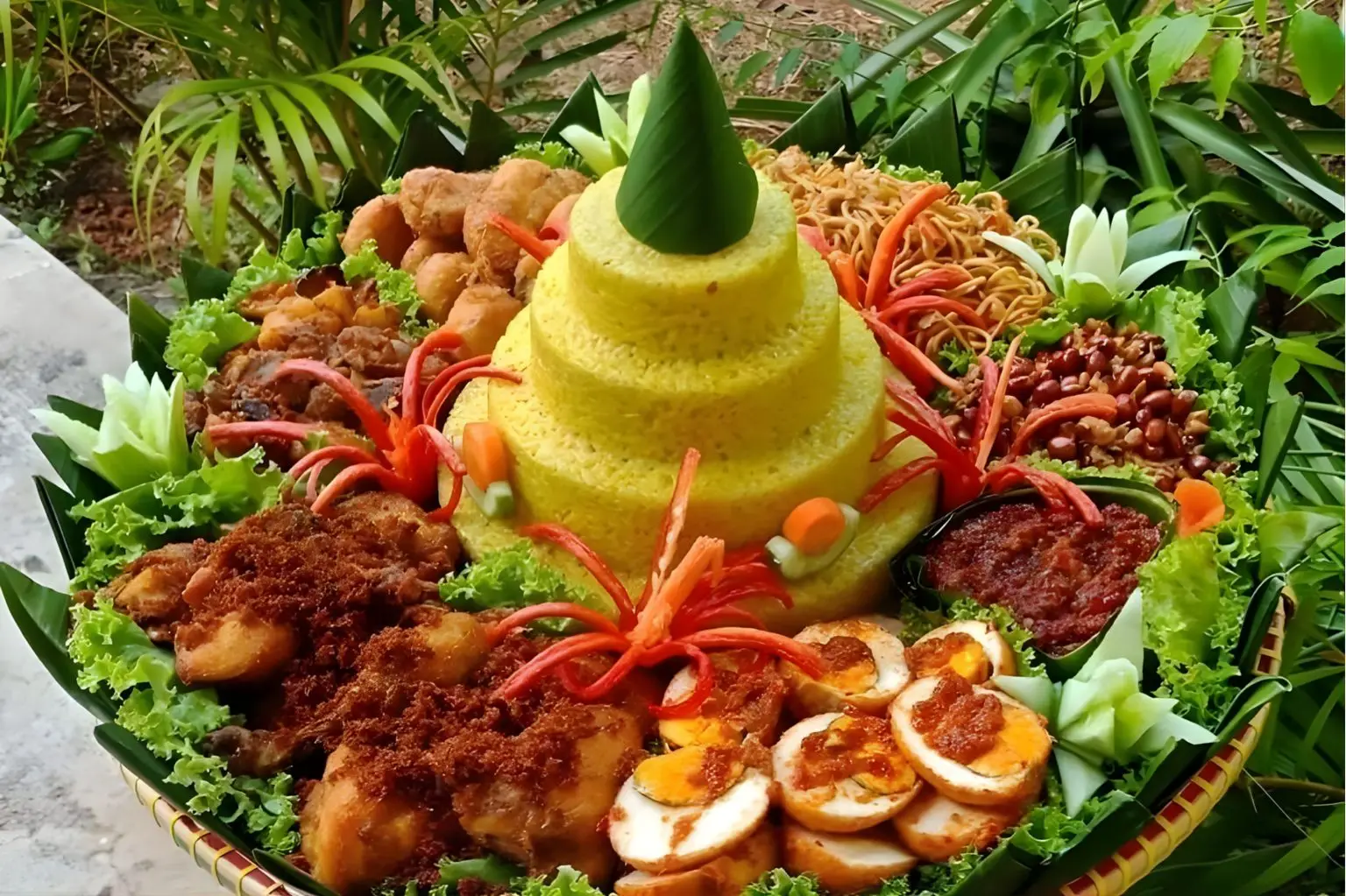 Nasi tumpeng kuning bertingkat dengan ayam goreng serundeng dan lauk pauk lengkap di atas tampah.