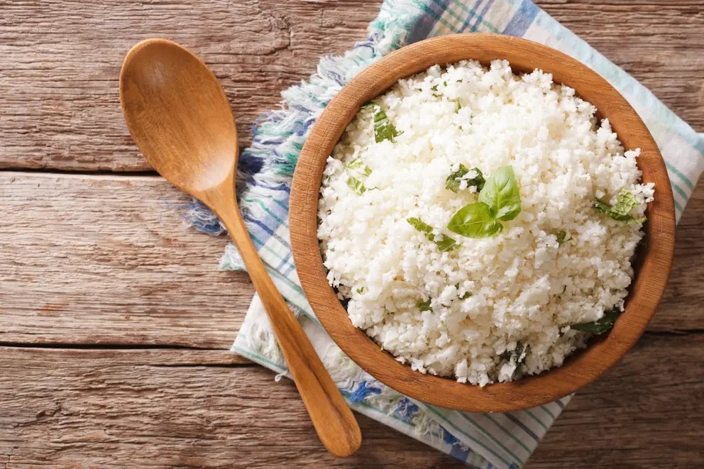 Tampilan top-down dari mangkuk kayu pedesaan berisi nasi kembang kol (cauliflower rice) parut, dihiasi daun kemangi segar dan parutan kulit jeruk. Sendok kayu diletakkan di samping di atas kain serbet dan meja kayu lapuk, sebagai alternatif pengganti nasi putih yang sehat.