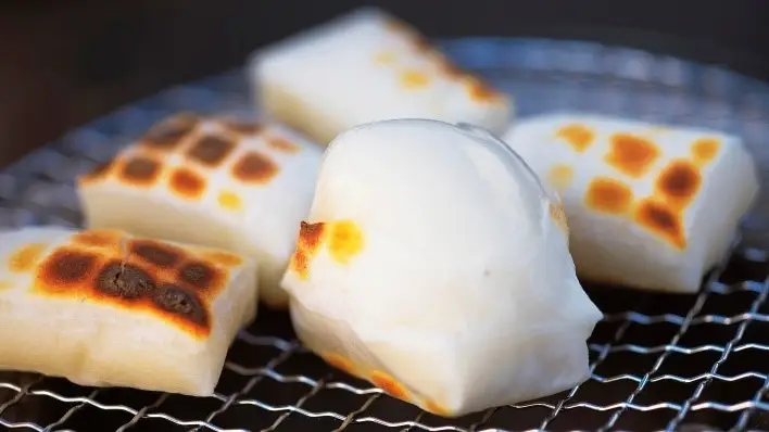 eberapa blok mochi putih (kirimochi) yang sedang dipanggang di atas kisi panggangan besi dengan bercak cokelat keemasan hasil karamelisasi.