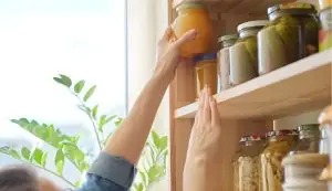 Tangan seseorang mencapai toples kaca berisi bumbu di rak kayu dapur yang rapi dengan tanaman di samping jendela cerah.